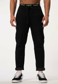 Schwarze Cargo-Hosen mit elastischem Taillenbund und Kordelzug, ausgestattet mit Seitentaschen, schmal zulaufenden Beinen und umgeschlagenen Bündchen. Getragen mit schwarzen Turnschuhen.
