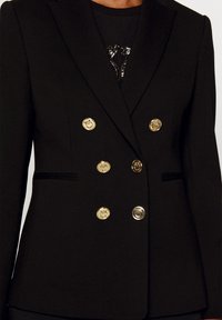 Black double-breasted blazer with gold round buttons, notched lapels, and subtle pockets. Smooth fabric with a tailored fit.