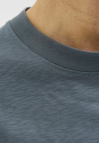 Grå bomulds T-shirt med struktureret finish, der har en ribbet rund halsudskæring og glat stof. Nærbillede fremhæver synningsdetaljen.