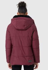 Persoon met een krullende bun, gekleed in een kastanjebruine gewatteerde jas met capuchon en zwarte bontdetails, gezien van achteren.