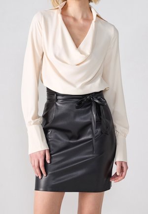 Vrouw die een crèmekleurige blouse met lange mouwen en een gedrapeerde halslijn draagt en een zwarte rok van namaakleer, gestrikt met een strik in de taille.