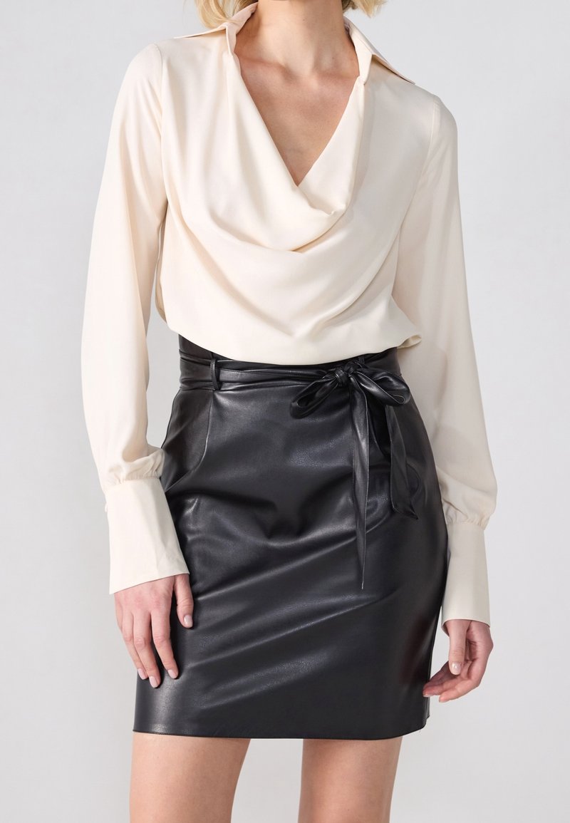Femme portant un chemisier crème à manches longues avec un col drapé et une jupe noire en simili cuir nouée d’un nœud à la taille.