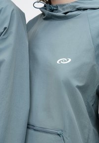 Lyseblå hættejakke med lynlåslomme foran og elastisk snøre ved halsen. Har en hvid logo på brystet. Silkeagtig tekstur.