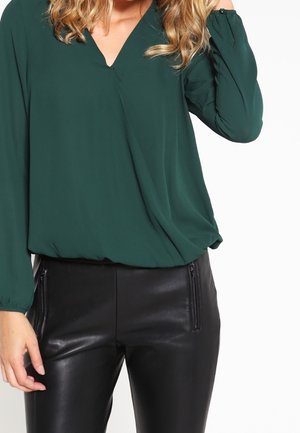 Femme portant un chemisier vert foncé à manches longues et un pantalon en cuir noir avec des poches zippées, posant avec une main près de ses cheveux.