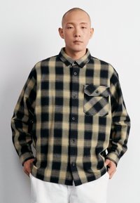 Camisa de franela a cuadros en negro y amarillo pálido con cuello de botón, bolsillo frontal y mangas largas, exhibida en un modelo sobre un fondo claro.