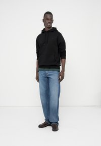 Zwarte hoodie met een voorzak, gedragen over een groen shirt; gecombineerd met lichtblauwe spijkerbroek en bruine lederen schoenen.