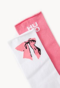 Cotton blend socks: one white with a pink bow graphic, the other solid pink with "HUGO" in white text. Smooth texture, ankle-length design.