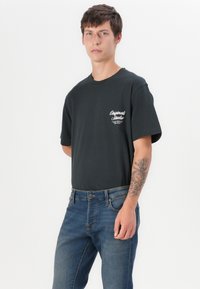 Camiseta de algodón negra con cuello redondo y mangas cortas, que presenta un logo blanco en el lado izquierdo del pecho. Llevada con jeans de mezclilla azul.