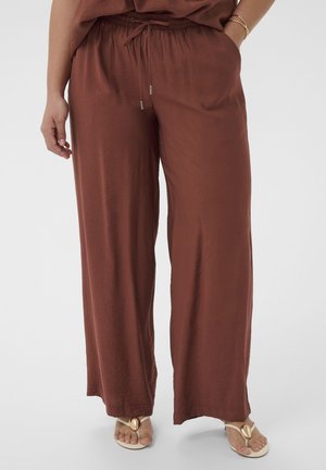 Persoon die losse bruine broek met aantrekkoord draagt, één hand in de zak en beige sandalen met gouden versieringen.