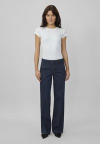 Een model staat centraal, gekleed in een aansluitend wit topje en een donkerblauwe wijde jeans, op een neutrale grijze achtergrond. Haar is los en natuurlijk.
