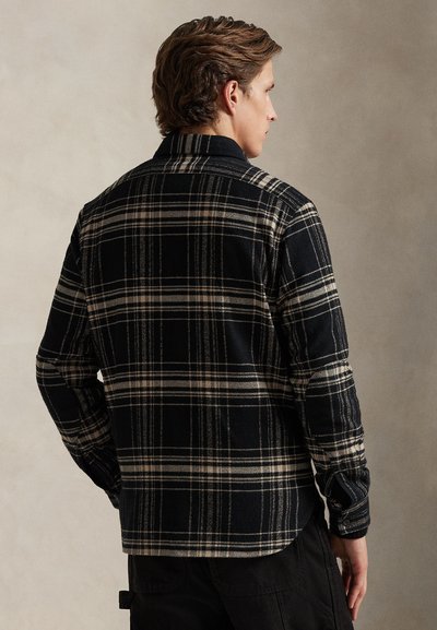Πουκάμισο από μάλλινο φανέλα με καρό μοτίβο σε μαύρο και μπεζ, με γιακά και μακριά μανίκια, χαρακτηριστικό απαλής υφής και κυρτή γραμμή στο κάτω μέρος, φορεμένο ανάποδα από μοντέλο.