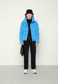 Doudoune bleue avec un col haut, des coutures contrastées et des boutons pression, associée à un col roulé noir et un pantalon noir. Sac noir texturé.