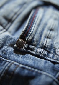 Zbliżenie na denimową tkaninę z metalowym guzikiem, czerwonym i niebieskim ściegiem oraz teksturowanym splotem. Skupienie na obszarze kieszeni i detalu zamka.