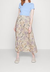 Helles blaues Kurzarmoberteil kombiniert mit einem beigen Maxirock im Blumenmuster, der graue, lila und orange Akzente aufweist. Dazu werden beige Slippers getragen.