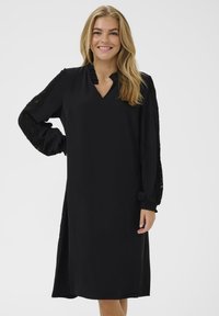 Vestido negro de tela suave, con escote en V, mangas largas con detalle de encaje y un diseño fluido hasta la rodilla. Simple y elegante.