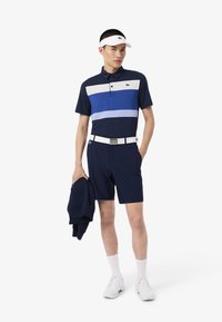 Polo shirt bleu marine avec des rayures horizontales bleues et blanches, associé à un short bleu marine. Comprend une visière blanche et un pull bleu marine drapé sur le bras.
