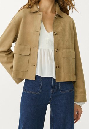 Woman wearing a tan suede buttoned jacket over a white textured blouse and high-waisted blue jeans with front patch pockets.
