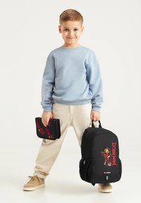 Zaino nero con design Ninjago, dettagli rossi e tasche laterali in rete. Bambino tiene un astuccio nero coordinato con elementi grafici rossi.