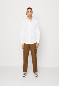 Camisa branca de botões com mangas longas, combinada com calças castanhas e ténis prateados. Modelo de pé, contra um fundo simples.