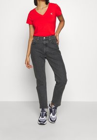 Červené tričko s V-výstřihem a logem CK, kombinované s černými džíny s vysokým pasem a modro-bílými teniskami. Jednoduchý design, volný střih.