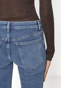 Pohled zezadu na osobu v nohavicích s vysokým pasem modré barvy a přiléhavém hnědém dlouhém rukávu, s jednou rukou odpočívající vedle boků.