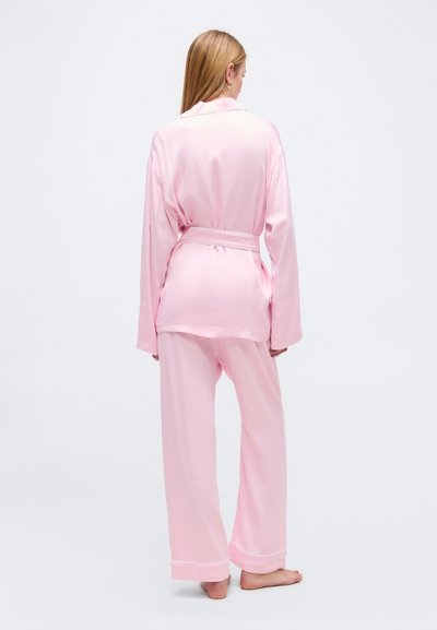 Γυναίκα που στέκεται ξυπόλητη, φοράει ανοιχτό ροζ σατέν πιτζάμα με μακριά μανίκια και παντελόνι με φαρδύ πόδι, φαινομένη από πίσω.