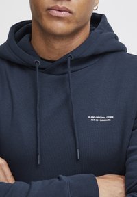 Hanorac bleumarin, realizat dintr-un material moale, cu un logo frontal în text alb. Include șnururi ajustabile și manșete cu nervuri.