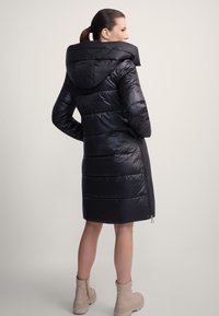 Kobieta ubrana w czarną, długą pikowaną kurtkę z dużym kapturem, ręce w kieszeniach, w beżowych botkach do kostki, stojąca na jednolitym tle.