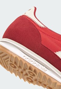 Červená športová obuv z brušenej kože a syntetických materiálov s krémovými akcentmi, s textúrovanou podrážkou a výrazným logom. Na päte je viditeľný detail vyšívania.