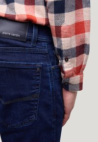 Donkerblauwe denim jeans met een subtiele, gestructureerde afwerking. Voorzien van een zwart leren label op de tailleband en zichtbare stiksels. Rood en marineblauw geruite shirtmouwen zijn gedeeltelijk zichtbaar.