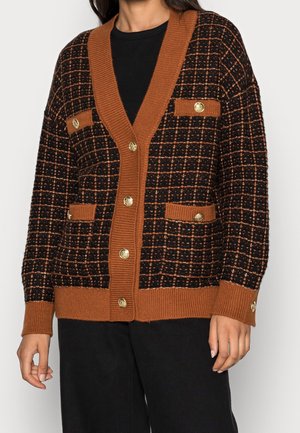 Cardigan a cuadros en negro y marrón con botones dorados, bordes acanalados y cuatro solapas de bolsillo, llevado sobre una blusa negra.