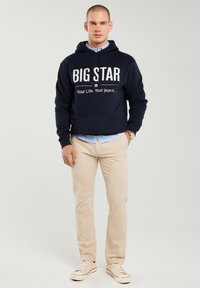 Vyriškis dėvi tamsiai mėlynį džemperį su užrašu „BIG STAR Your Life. Your Jeans.“, smėlio spalvos kelnes, mėlyną marškinius ir baltus sportbačius, stovintis prie šviesaus fono.
