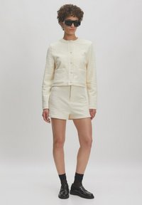 Lichtcrème, getextureerde twee-delige outfit met een cropped jas met knopen en zakken, gecombineerd met bijpassende shorts. Draag het met zwarte schoenen.