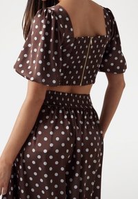 Brązowy top crop w kropki, z bufiastymi rękawami i kwadratowym dekoltem. Posiada marszczony tył oraz metalowy zamek błyskawiczny. Gładka faktura materiału.