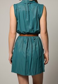 Short sleeveless teal dress with a collar, gathered waist, and brown belt. The fabric appears smooth and slightly glossy with a flowing silhouette.