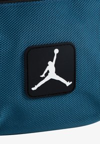 Tyrkis teksturert stoffveske med et fremtredende svart og hvitt Jordan-logo-patch, som har en silhuett av en figur som hopper med en basketball.
