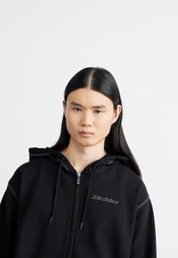 Crna zip-up dukserica s teksturiranom tkaninom, vezicama i malim sivim Dickies logotipom na prsima. Jednostavan, minimalistički dizajn.