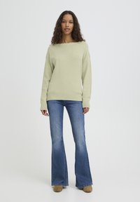 Pull tricot vert clair à large encolure, associé à un jean évasé bleu et des chaussures beige clair. La texture semble douce avec des poignets côtelés.