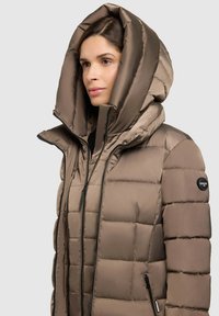 Ruda pūkinė striukė su didele, struktūrizuota kapuci, su užtrauktuku, juoda įranga ir logotipu ant rankovės.