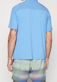 Man som bär en ljusblå kortärmad skjorta och shorts med flerfärgade pastellmönster, sedd bakifrån.