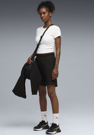 Woman with curly hair wearing white crop top, black shorts, black sneakers, white Puma socks, holding a black jacket with strap bag across shoulder.