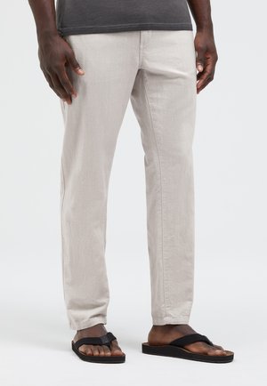 Ein Mann trägt hellbeige Hosen, schwarze Flip-Flops und ein dunkelgraues Hemd und steht vor einem schlichten, hellen Hintergrund.