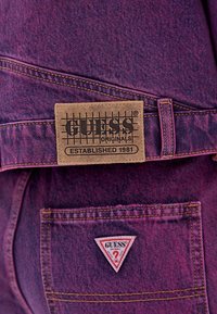 Paarse denim stof met gele stiksels; een bruin leren label met de tekst "GUESS" en een rasterpatroon. Bevat een rode driehoekige tag op de zak.