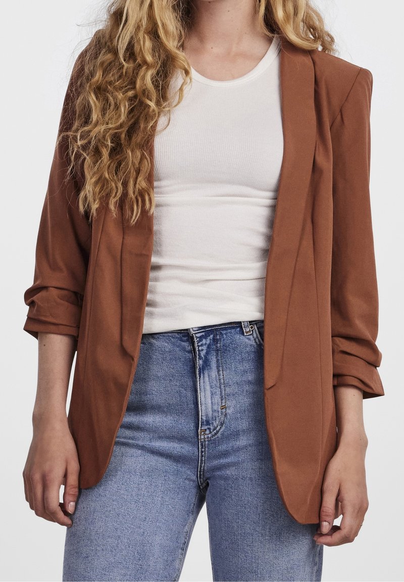 Blazer marron avec une coupe décontractée, manches longues retroussées, en tissu lisse. Porté par-dessus un haut blanc ajusté et associé à un jean bleu clair.