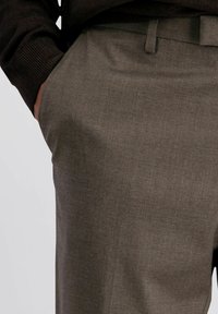 Persoon in bruine op maat gemaakte broeken met een hand nonchalant in de zijzak, gecombineerd met een donkere ribbeltrui.