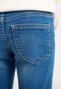 Detailný záber na zadnú vrecko modrých denimových nohavíc s oranžovým prešívaním, nosených s bielym rebrovaným topom.