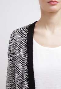 Černobílý pletený cardigan s otevřeným předním dílem, vybavený strukturovaným vzorem a hladkým černým lemováním kolem okrajů. Bílé tričko pod ním.