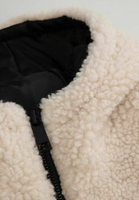 Kurtka polarowa w kolorze kremowym z miękką, teksturowaną powierzchnią, czarną podszewką i zapięciem na zamek z przodu.