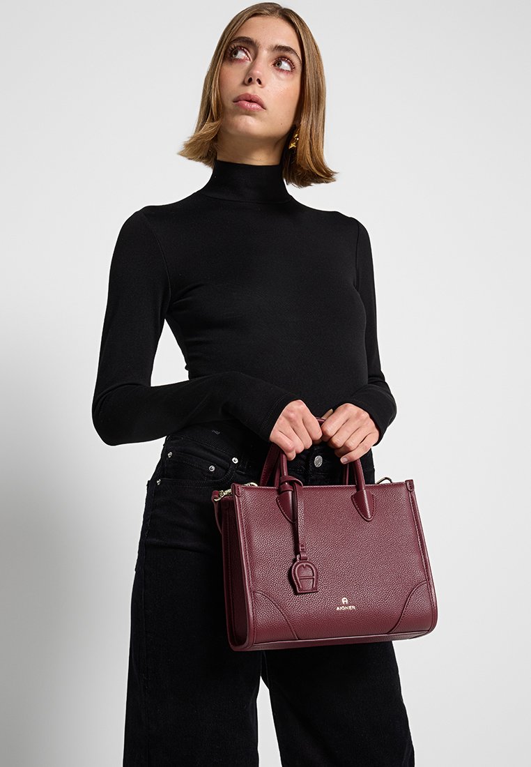 Burgunderfarbene Lederhandtasche mit strukturierter Oberfläche, klarer Form, doppelten oberen Griffen und goldenen Beschlägen. Dekorativer Anhänger am Griff befestigt.