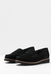 Melnas zamšādas loafers ar izšūtu dizainu, zemu papēdi un kontrastējošu brūnu zoli. Augšdaļai ir klasiskā mokasīnu forma ar krokām.